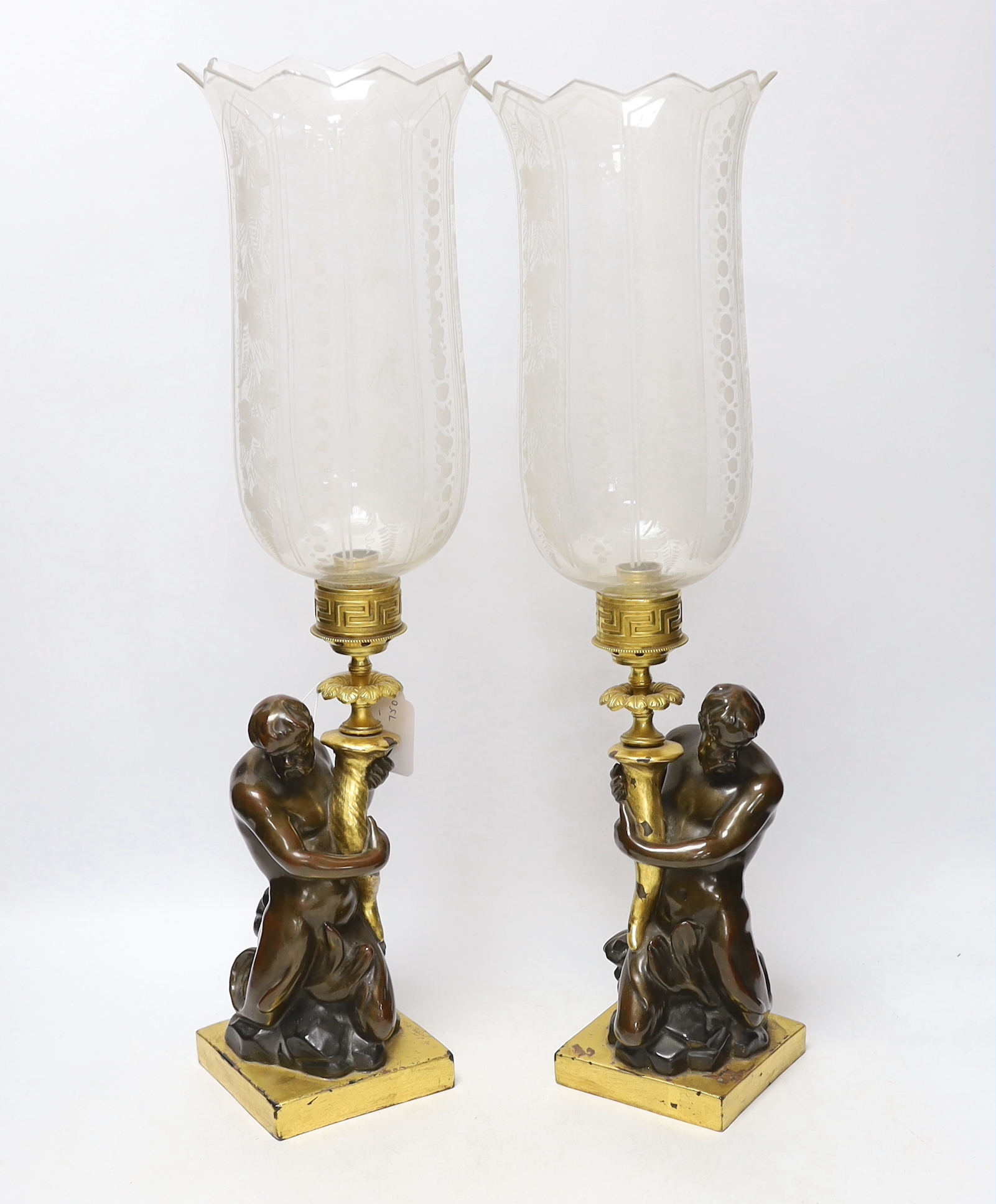 A pair of 19th century bronzed stoneware candlesticks, after a design by John Flaxman, each modelled as a triton holding a cornucopia, supporting a gilt brass sconce and engraved glass hurricane shade, 54cm high